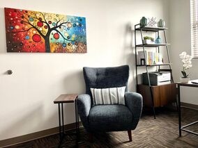 Dark blue chair, colorful tree painting, and books and plants seen on the shelf inside Dr. Penela’s therapy office in Pembroke Pines, Florida. 