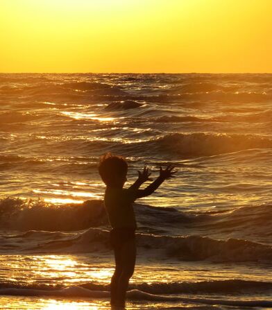 Child on beach shoreline with hands in air and sun setting in background.